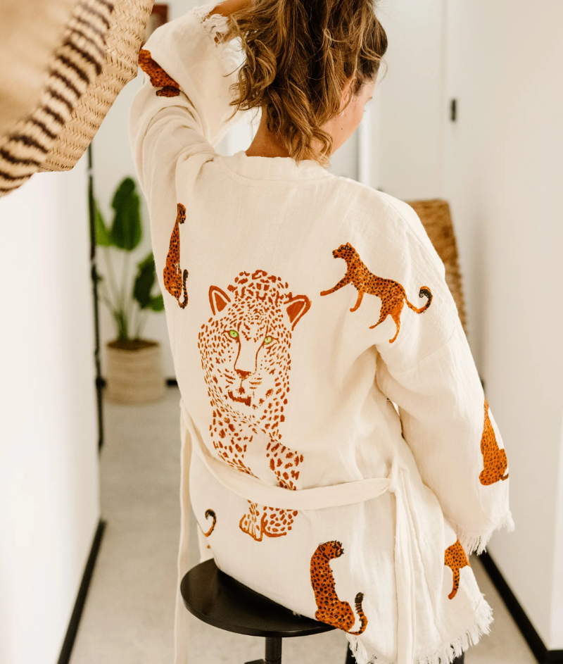 A woman viewed from behind wearing a cream-colored cotton robe with embroidered leopard designs in brown and orange, sitting on a black stool in a bright indoor setting.